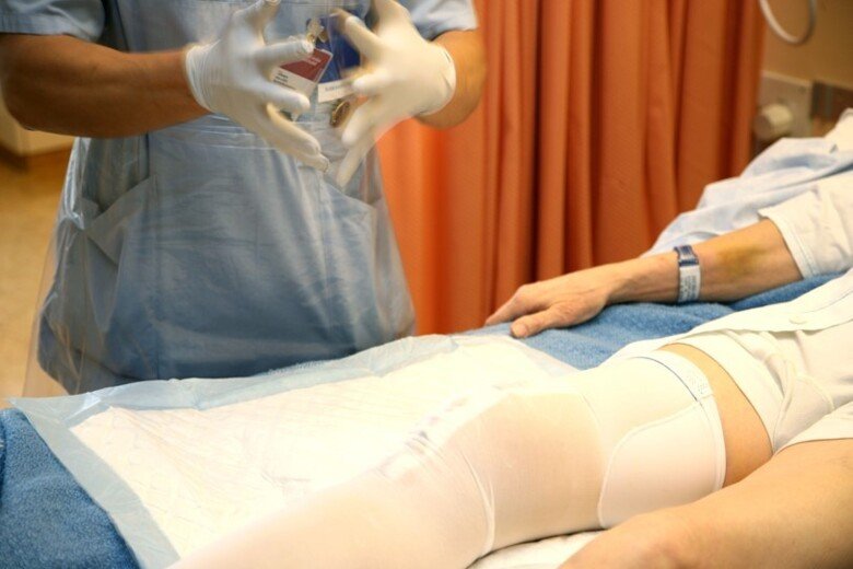 Bild på en persons händer vid en sjukhussäng där man ser en patients ben med förband. Illustrerar verksamhetsförlagd utbildning, fysioterapeutprogrammet.