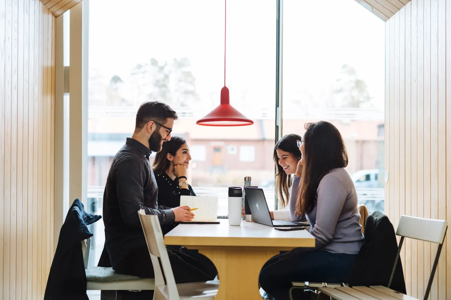 Fyra studenter sitter runt ett bord och samtalar. || Fyra studenter som sitter vid ett bord och studerar med sina laptops.