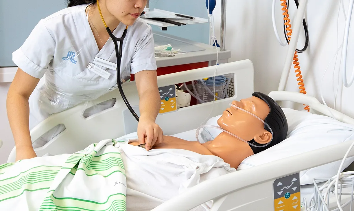 En läkarstudent står lutad över en sjukhussäng där en docka ligger.