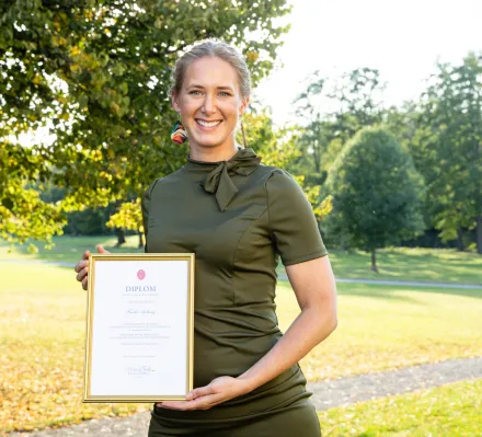 En bild på Frida Axberg den en ljushårig ung kvinna i grön klänning som står framför ett fönster och ler mot kameran.