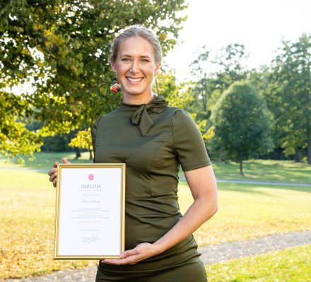 En bild på Frida Axberg den en ljushårig ung kvinna i grön klänning som står framför ett fönster och ler mot kameran.